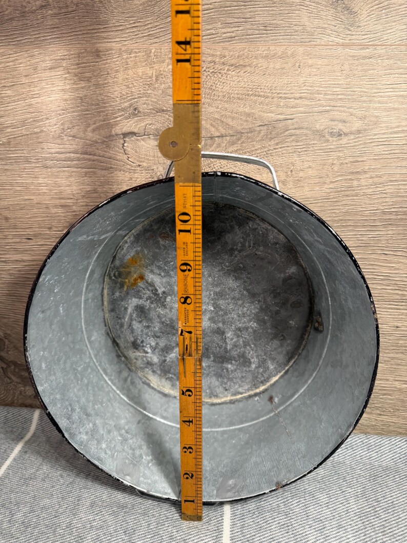 Op de afbeelding: Een ronde gegalvaniseerde metalen bak met een zwarte rand en een zilverkleurig metalen handvat. Een gele opvouwbare houten liniaal is verticaal in het midden geplaatst en toont een diameter van ongeveer 26,5 centimeter voor de opening.