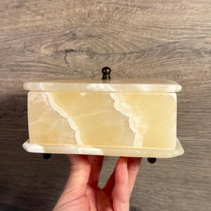 Peut inclure: Une boîte rectangulaire en onyx jaune clair avec un couvercle à charnière et un petit bouton rond en laiton. La boîte présente un motif marbré blanc et repose sur quatre petits pieds sombres. La boîte est tenue dans une main.