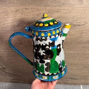 Puede incluir: Tetera de metal pintada a mano con diseño de vaca. La tetera tiene un asa y tapa azules, con detalles amarillos y verdes. El cuerpo es blanco con patrones de vaca negros y verdes.