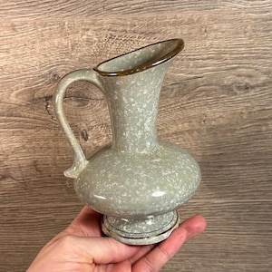 May include: A small, gray ceramic pitcher with a speckled glaze and a gold rim. The pitcher has a rounded body and a short, flared spout. It has a single handle and a small, round base.
