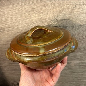 May include: A brown ceramic casserole dish with a lid and a handle. The dish has a mottled glaze with brown, green, and white tones.