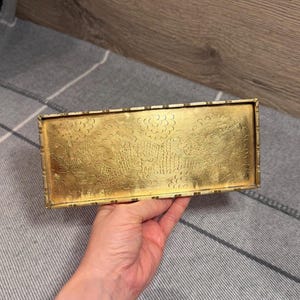 Puede incluir: Una bandeja rectangular de metal color dorado con un diseño grabado detallado de un dragón entre nubes estilizadas. La bandeja tiene un borde elevado con un patrón de muescas repetitivo a lo largo de los bordes, con acabado metálico.
