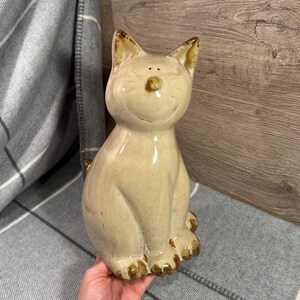 Könnte beinhalten: Eine Keramik-Katzenfigur in sitzender Position. Die Katze hat eine hellbeige Farbe mit dunkelbraunen Akzenten an Ohren, Nase und Pfoten. Sie hat einen lächelnden Ausdruck und ist etwa 18 cm hoch.