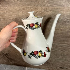 May include: White ceramic coffee pot with a long spout and a handle. The pot is decorated with a colorful fruit pattern featuring apples, pears, and berries. The lid also has a fruit design.
