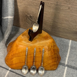 May include: Four silver teaspoons with ornate handles displayed on a wooden stand. The spoons are arranged in a row, with the handles pointing upwards. The stand is made of dark wood and has a simple design.