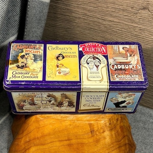 Op de afbeelding: Een vintage blik met een paarse achtergrond en kleurrijke afbeeldingen van Cadbury's chocoladekoekjes. De doos bevat verschillende Cadbury's-producten, waaronder Cadbury's Pure Milk Chocolate, Cadbury's Chocolate Biscuits en Cadbury's Dairy Milk Chocolate Biscuits.