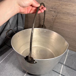 Puede incluir: Un caldero de aluminio vintage con un asa de metal curvada. El caldero tiene una boca ancha y abierta y un cuerpo redondeado. El asa está sujeta a los lados del caldero con soportes metálicos. El caldero es sostenido por una persona.