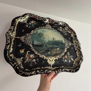 Könnte beinhalten: Schwarzes Dekotablett mit goldenem Rand und Landschaftsmalerei in der Mitte. Das Tablett hat einen gewellten Rand und ist mit Blumen- und Schmetterlingsmotiven verziert. Misst etwa 40 cm.