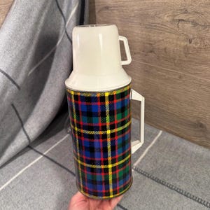 Puede incluir: Termo vintage con tapa y asa blancas, y un colorido estampado de cuadros en azul, verde, rojo y amarillo. El termo tiene un asa blanca y una taza blanca a juego que sirve de tapa.