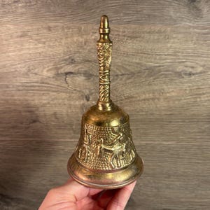 May include: A gold-colored bell with an ornate handle and a detailed design of figures on the body.