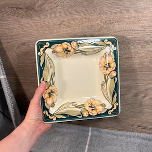Puede incluir: Un plato cuadrado de cerámica con un diseño floral verde y blanco. El plato tiene un fondo blanco con un borde verde y flores amarillas con acentos rosados.
