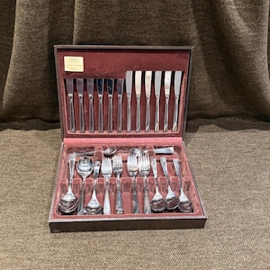 May include: A silver-toned stainless steel flatware set housed in a dark brown storage case with a maroon velvet interior. The set includes knives, forks, and spoons arranged in molded slots. A small rectangular label reads MIMOSA.