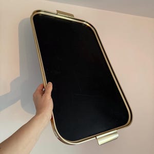 Puede incluir: Una bandeja rectangular con interior negro y marco de metal dorado. La bandeja tiene esquinas redondeadas y dos asas doradas. La bandeja se sostiene contra una pared de color rosa claro.