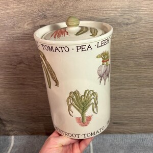 May include: A white ceramic kitchen canister with a lid, featuring illustrations of vegetables like tomatoes, peas, and leeks. The canister has a cylindrical shape and is held by a person's hand.