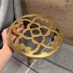 May include: A brass trivet with a decorative openwork design. The trivet has a circular shape with a floral pattern and three wooden legs. The brass has a warm, golden hue. This item is designed to protect surfaces from hot cookware.