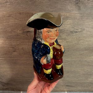 Könnte beinhalten: Ein Keramikkrug in Form eines Mannes mit Dreispitz und einem kleinen Krug. Die Figur trägt einen blauen Mantel, eine gelbe Weste, rote Kniebundhosen und schwarze Schuhe. Der Krug hat einen braunen Sockel und einen Griff auf der Rückseite.