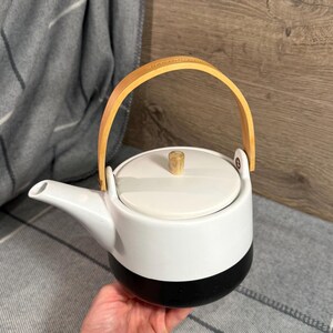 May include: A two-toned teapot featuring a matte white upper section and a solid black base. The teapot includes a matching white lid with a small wooden knob handle and a curved light-coloured wooden swing handle attached to the sides.