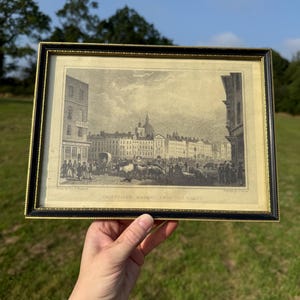 May include: Framed black and gold print of a historical cityscape, featuring a bustling market scene with buildings, people, and livestock. The print is labeled "Smithfield Market, From the East."