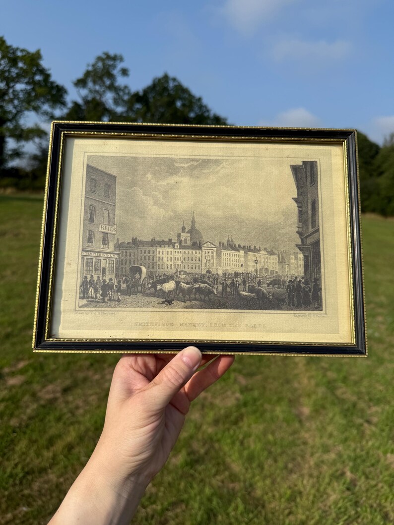 May include: Framed black and gold print of a historical cityscape, featuring a bustling market scene with buildings, people, and livestock. The print is labeled "Smithfield Market, From the East."