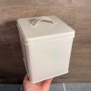 Może przedstawiać: Kwadratowy, kremowy metalowy pojemnik do przechowywania z zakrzywionym uchwytem na pokrywie. Pojemnik ma prosty, czysty design i prawdopodobnie służy do przechowywania przedmiotów. Pojemnik jest trzymany przez osobę.