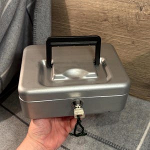 Puede incluir: Una caja de metal plateado con un asa negra y cerradura de llave. La caja es rectangular con esquinas redondeadas y una parte superior ligeramente abovedada. La llave está en la cerradura con una cuerda verde.