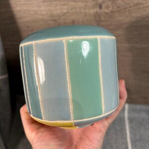 Op de afbeelding: Een keramische plantenpot met een gestreept ontwerp in tinten blauw en groen. De pot heeft een gele rand.