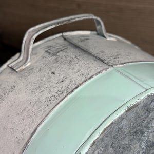 Op de afbeelding: Een ronde metalen container met een gebogen handvat aan de bovenkant. Het oppervlak toont een combinatie van verweerd, getextureerd grijs gegalvaniseerd metaal en gladde, mintgroen geschilderde secties met zichtbare oxidatie aan de randen.