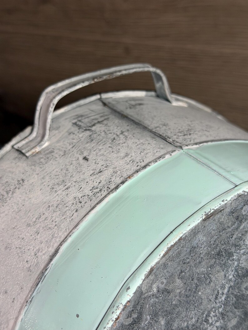 Op de afbeelding: Een ronde metalen container met een gebogen handvat aan de bovenkant. Het oppervlak toont een combinatie van verweerd, getextureerd grijs gegalvaniseerd metaal en gladde, mintgroen geschilderde secties met zichtbare oxidatie aan de randen.