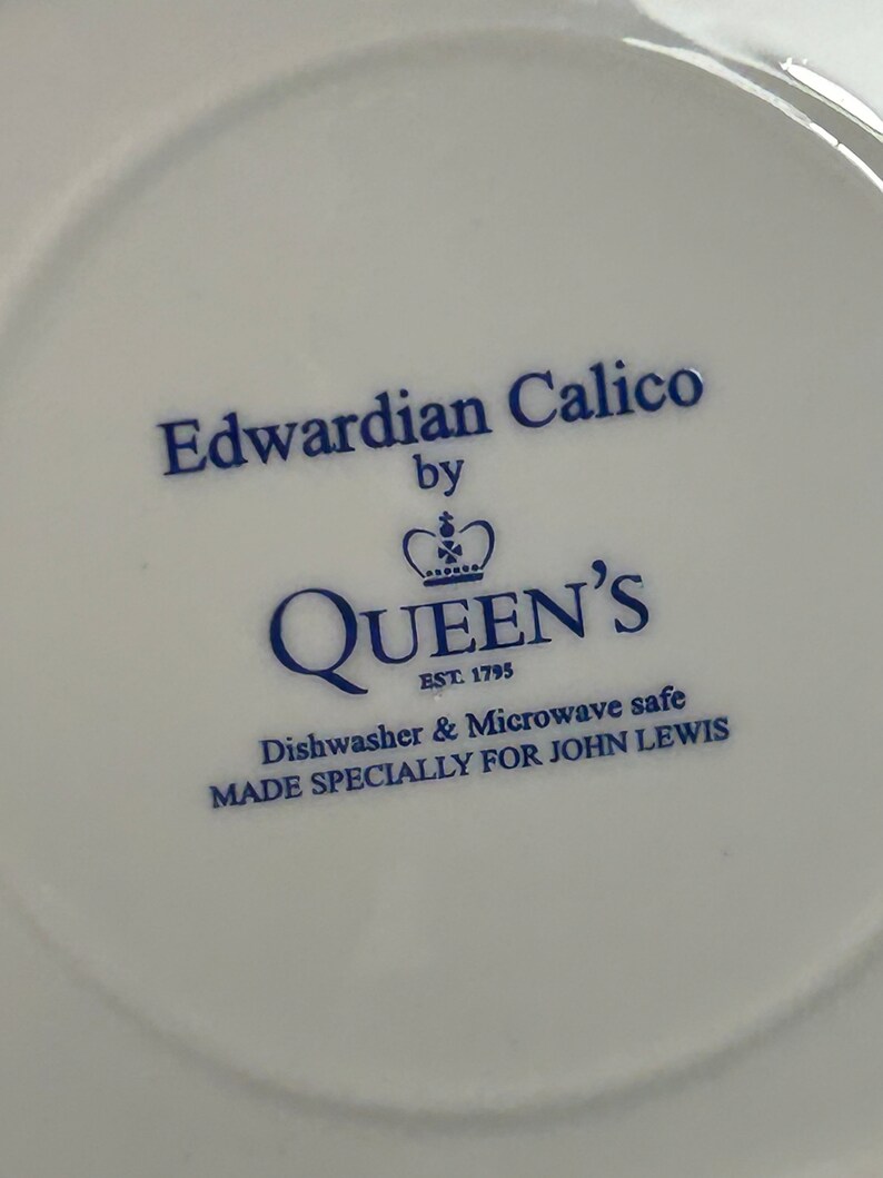 May include: A white ceramic saucer with blue text that reads "Edwardian Calico by QUEEN'S EST 1795 Dishwasher & Microwave safe MADE SPECIALLY FOR JOHN LEWIS".