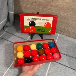 May include: A set of Guinness Brothers Snooker & Pool balls in a red cardboard box. The box lid features the text GUINNESS BROTHERS SNOOKER SPECIALISTS LTD and SNOOKER & POOL BALLS with illustrations of balls. The box contains twenty-two colorful balls.