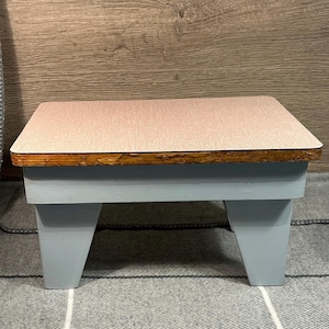 Puede incluir: Un pequeño taburete de madera azul claro con una parte superior marrón claro. La parte superior tiene una superficie texturizada.