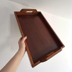 Puede incluir: Una bandeja de madera rectangular con un acabado marrón oscuro. La bandeja tiene dos asas recortadas y se sostiene en ángulo contra un fondo blanco. La veta de la madera es visible en la superficie.