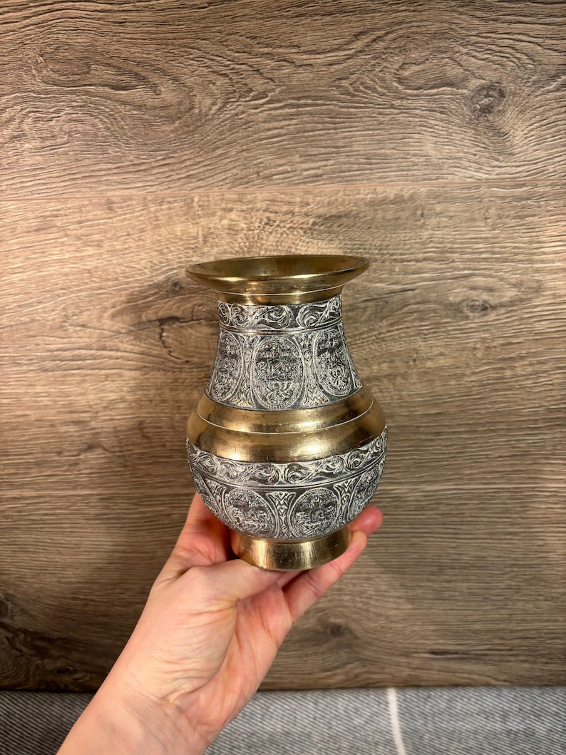 K&ouml;nnte beinhalten: Eine kleine, goldfarbene Vase mit aufwendigen, silberfarbenen, ge&auml;tzten Designs. Die Vase hat einen runden K&ouml;rper und einen schmalen Hals.