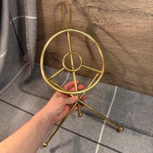 May include: A gold-colored metal magazine rack with a circular design and three legs. The rack has a handle at the top and a small circular detail in the center. The legs have rounded tips.
