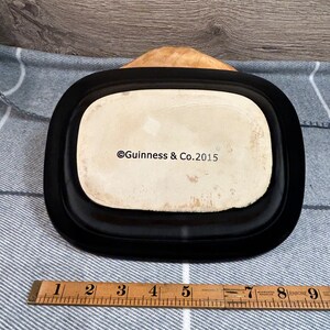 May include: A black rectangular dish with rounded corners, featuring a cream-colored interior. The text "Guinness & Co. 2015" is visible. A wooden ruler with inch markings is placed near the dish.