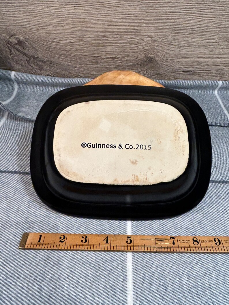 May include: A black rectangular dish with rounded corners, featuring a cream-colored interior. The text "Guinness & Co. 2015" is visible. A wooden ruler with inch markings is placed near the dish.
