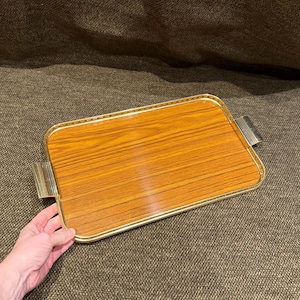 May include: A rectangular serving tray featuring a wood grain pattern surface, enclosed by a polished gold-tone metal frame with a decorative dotted border and two integrated gold-tone metal handles on the short sides of the tray.