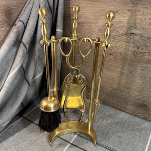 May include: A brass fireplace tool set with a stand. The set includes a brush with black bristles, tongs, a shovel, and a poker. The tools have decorative handles and are arranged on a curved base. The set is made of brass.