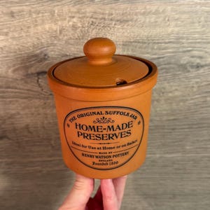 May include: A tan-colored Suffolk jar with a lid and a knob. The jar has a label that reads "HOME-MADE PRESERVES" and "HENRY WATSON POTTERY ENGLAND Founded 1800". The jar is held by a person.