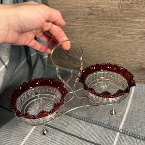 May include: A vintage double dish with a silver handle and base. The two clear glass bowls have a decorative red rim. The dish is designed with a silver handle for easy carrying and is supported by small silver feet.