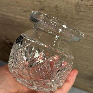 Op de afbeelding: Transparante kristallen pot met een brede opening en een decoratief geslepen patroon. De pot heeft een afgeronde vorm en wordt in een hand gehouden. Het kristal reflecteert licht en toont het ingewikkelde ontwerp. De pot is waarschijnlijk voor decoratieve of opslagdoeleinden.
