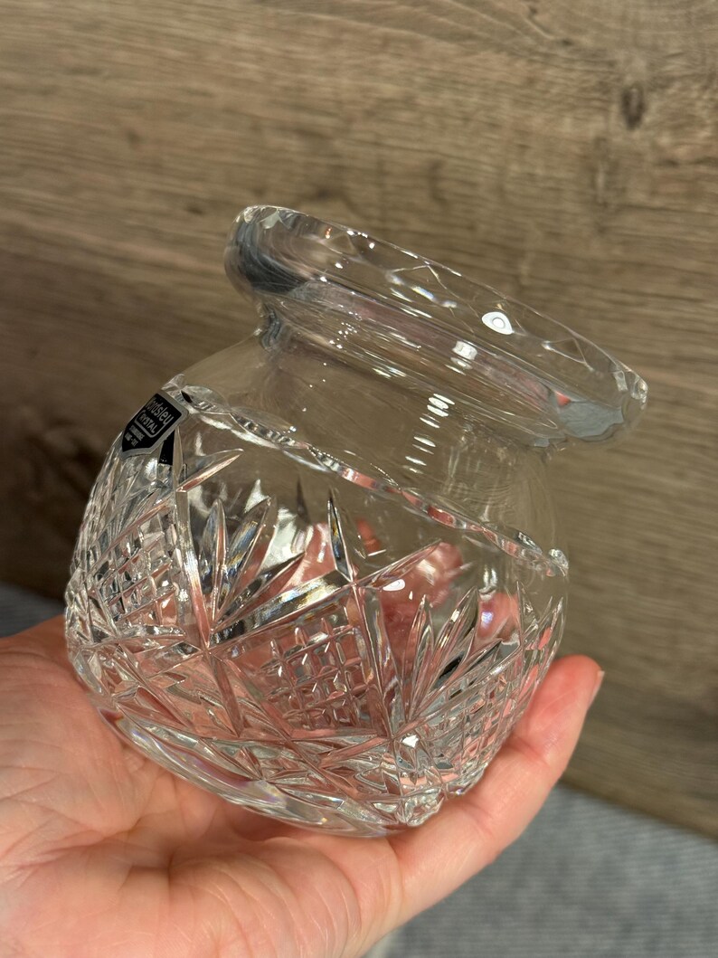 Op de afbeelding: Transparante kristallen pot met een brede opening en een decoratief geslepen patroon. De pot heeft een afgeronde vorm en wordt in een hand gehouden. Het kristal reflecteert licht en toont het ingewikkelde ontwerp. De pot is waarschijnlijk voor decoratieve of opslagdoeleinden.