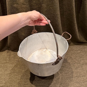 May include: A vintage silver-toned metal pot featuring a rounded basin, a pouring spout on one side, and a curved wire handle attached to two metal brackets. The interior shows signs of wear and discolouration, and the exterior has a matte finish.