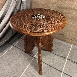 May include: A small, round wooden table with detailed floral carvings. The table features three ornate legs and an inlaid decorative centre. The wood has a warm, brown tone, and the table is approximately 46 cm in diameter.