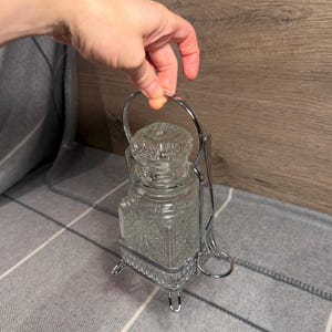 Puede incluir: Un tarro de cristal transparente con un patrón de diamantes texturizado y una tapa de cristal a juego, alojado en un marco de metal plateado con cuatro patas pequeñas, un asa curva y un soporte lateral para un pequeño utensilio de servicio.