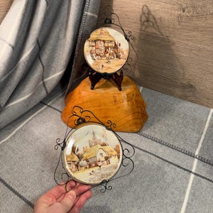 May include: Two decorative plates with black metal frames. The plates feature vintage illustrations of English cottages. One plate is on a wooden stand, the other is being held by a hand.