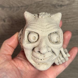 May include: A light beige, sculpted face with horns, large eyes, and a wide grin. The detailed sculpture has a textured surface and is held in a hand. The face appears to be a decorative item or figurine.