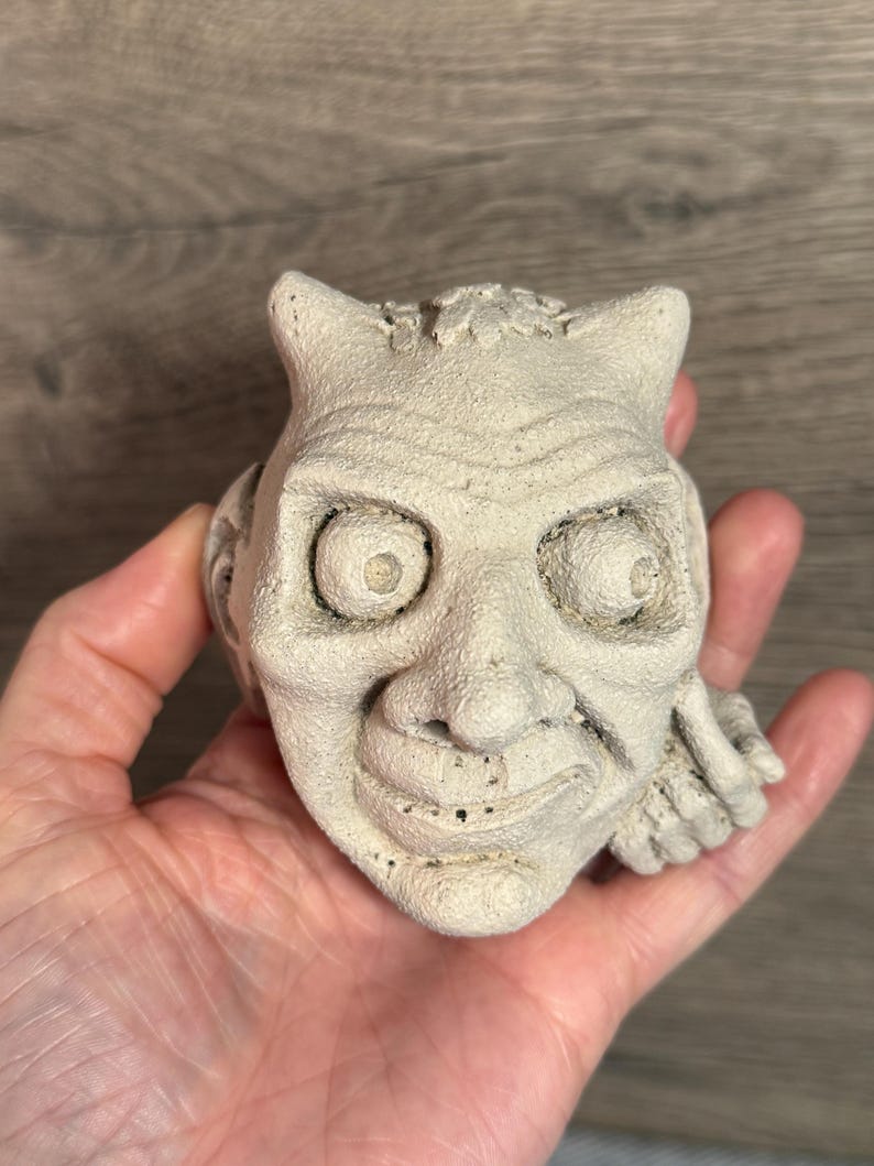 May include: A light beige, sculpted face with horns, large eyes, and a wide grin. The detailed sculpture has a textured surface and is held in a hand. The face appears to be a decorative item or figurine.