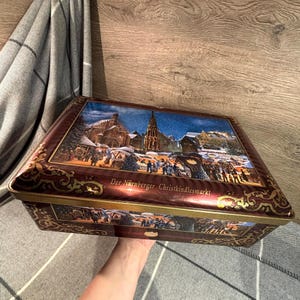 Puede incluir: Una lata rectangular de color granate con una ilustración detallada de una escena invernal. La tapa presenta una plaza nevada con una iglesia, puestos de mercado y gente. El texto "Der Nürnberger Christkindlesmarkt" es visible.