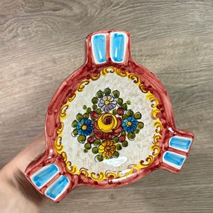 May include: A red and white ceramic ashtray with a floral design. The ashtray has three sections and is decorated with blue and yellow accents.